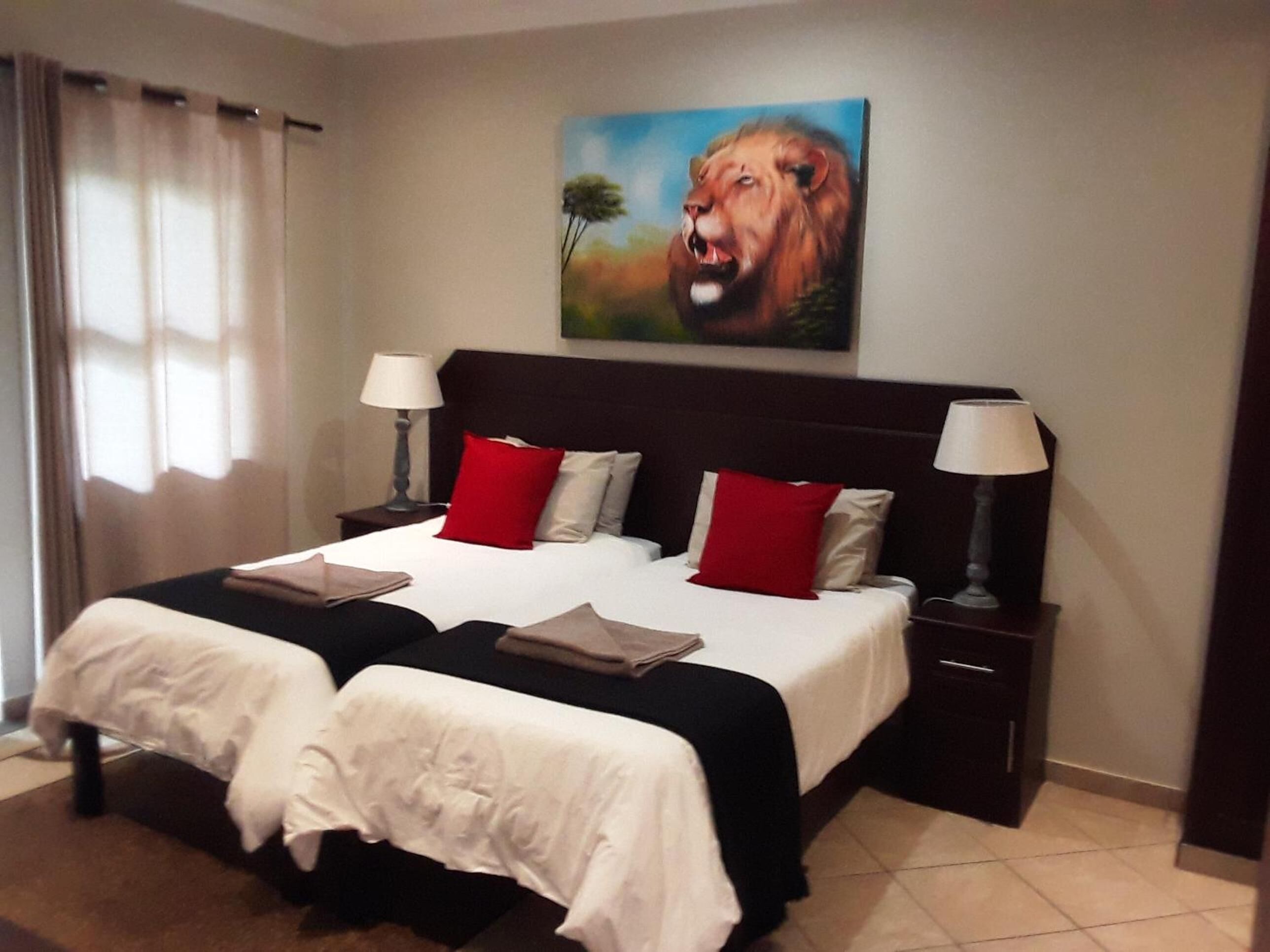 Una cama doble con ropa de cama blanca y roja, dos mesitas de noche con lámparas, y un cuadro de un león en la pared.