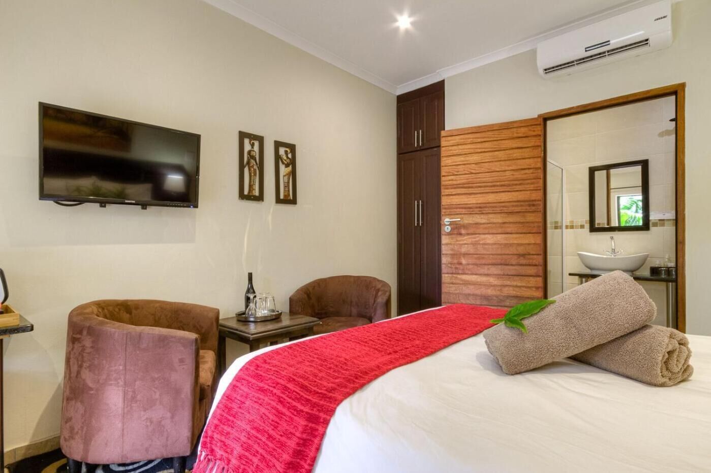 Habitación de hotel con cama, dos sillas, televisor, armario de madera y baño con lavamanos y espejo.