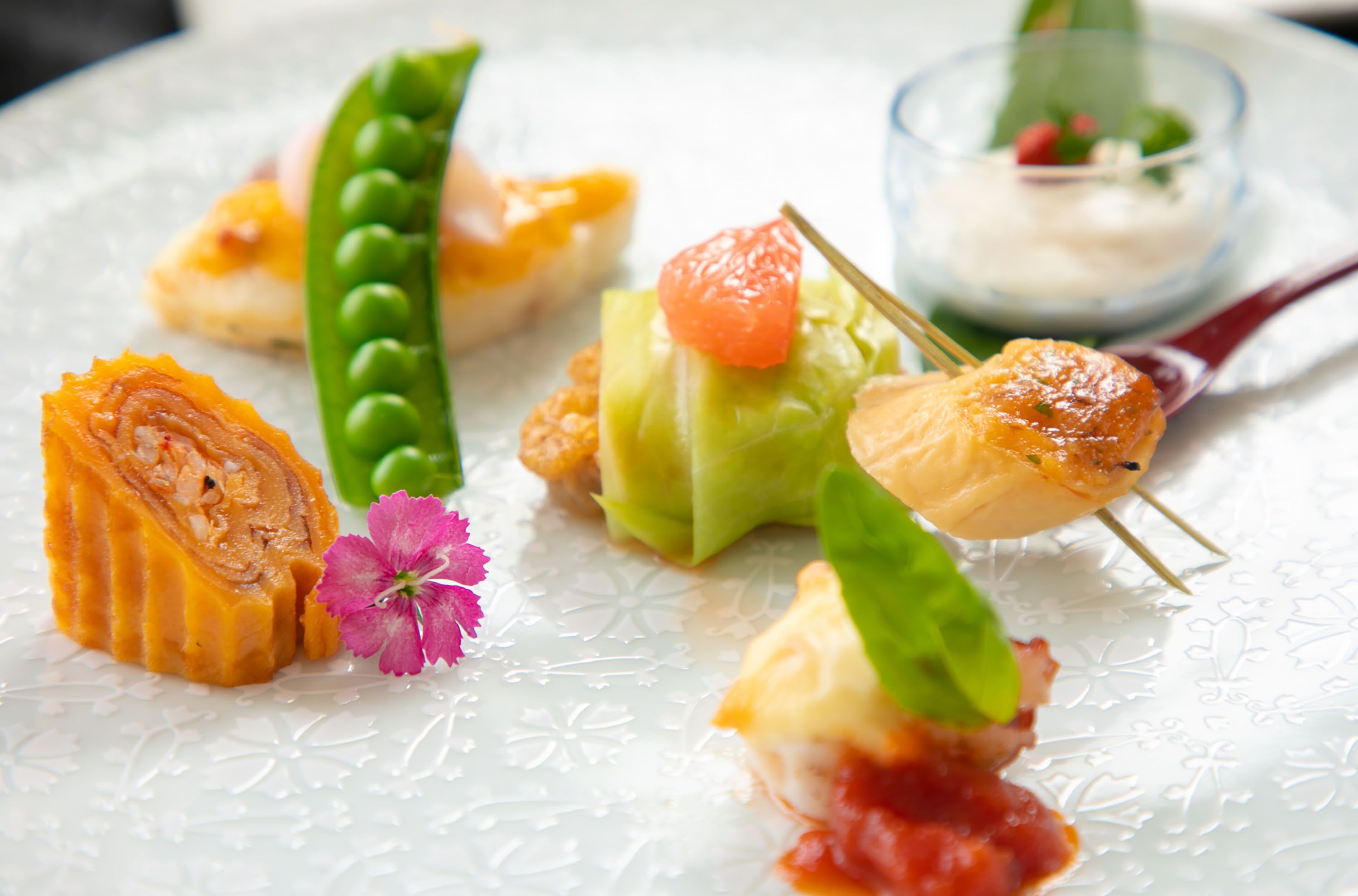 A plate with various food items including a rolled pastry, a pea pod, a carrot, and a skewer with a piece of meat and a cherry tomato.