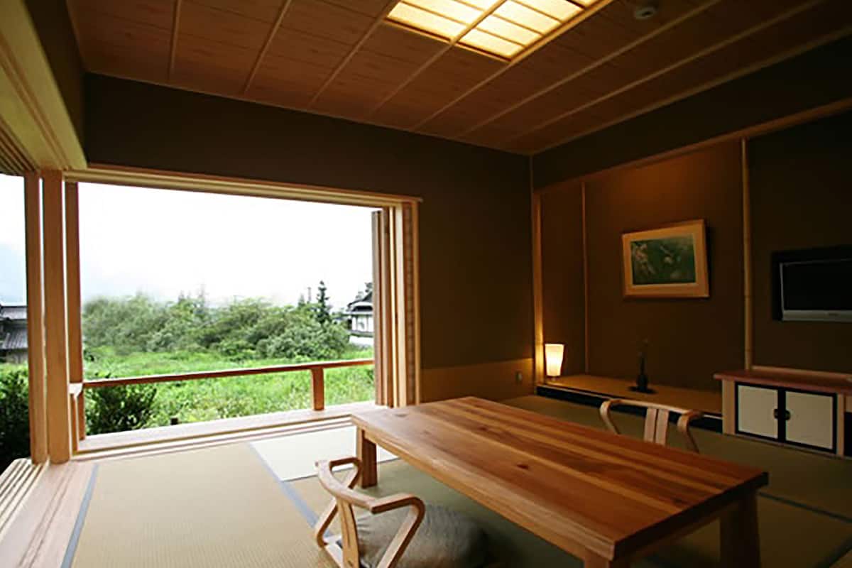 Una habitación tradicional japonesa con suelo de tatami, una mesa de madera baja y vistas a la vegetación exterior.