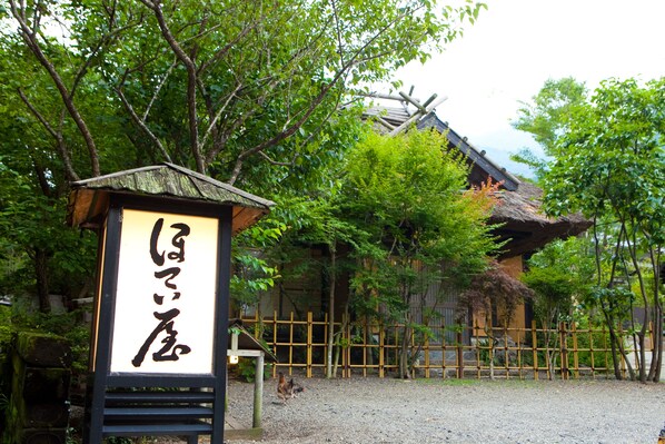 Un edificio japonés tradicional con techo de paja, rodeado de una valla de madera y abundante vegetación.