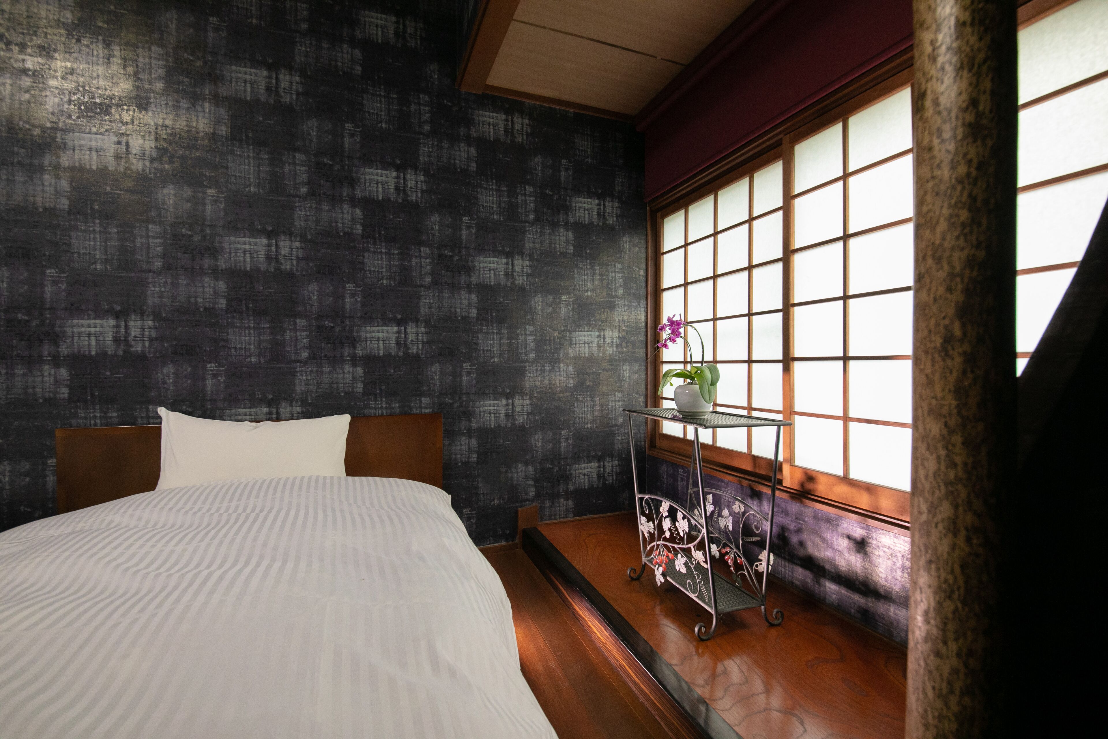 Un dormitorio con cama, pared oscura con textura, ventana con vistas y una pequeña mesa con una planta.