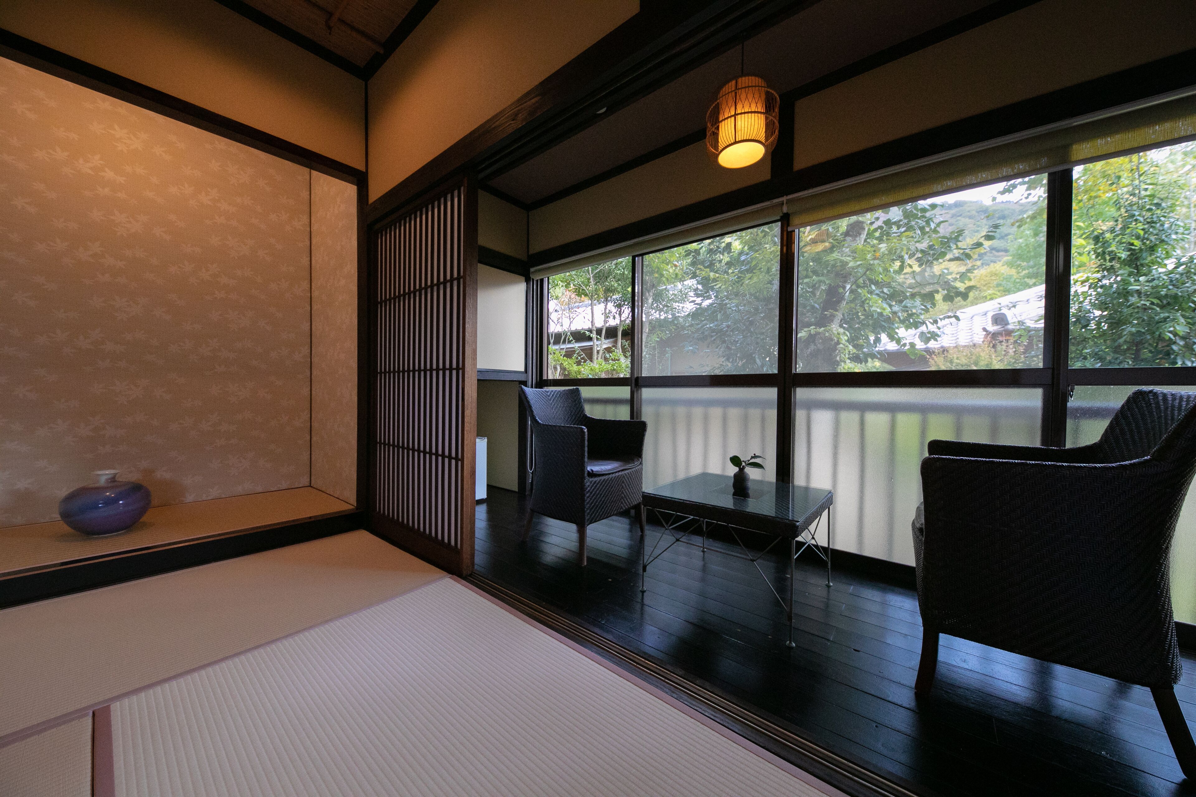 Een traditionele Japanse kamer met tatami vloerbedekking, een lage tafel met een plant, een stoel en uitzicht op groen buiten.