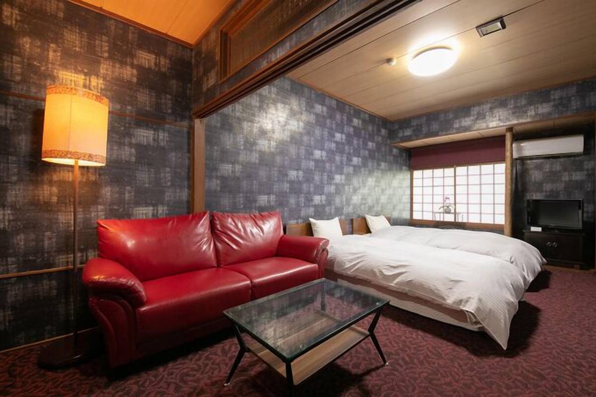 Una habitación de hotel con un sofá de cuero rojo, una mesa de centro de vidrio, una cama con ropa blanca, un televisor y un ventanal con vistas.