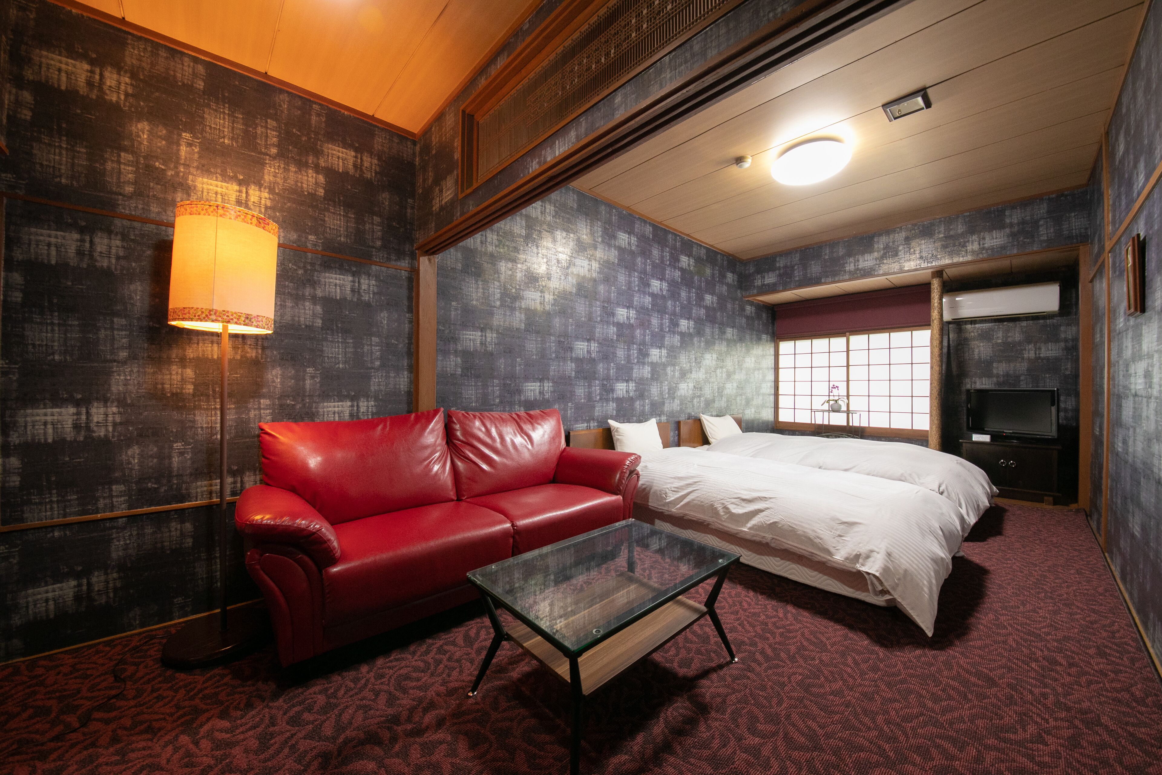 A hotel room with a red leather sofa, a glass coffee table, a white bed, and a television.