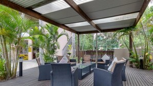 Un área de estar al aire libre con mobiliario de mimbre y una mesa con cubierta de vidrio, rodeada de abundante vegetación y una terraza de madera.