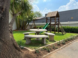 Une aire de jeux avec un toboggan et une structure d’escalade, entourée d’une pelouse bien entretenue et d’un arbre.