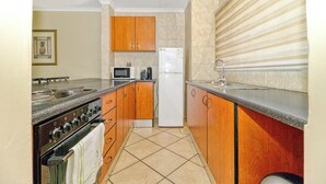 A compact kitchen with wooden cabinets, a white refrigerator, a microwave, and a stove. - Bm Gardens Apartment Hotel (Germiston)