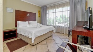 A hotel room with a bed, a desk with a television, a window with curtains, and a wall-mounted air conditioning unit. - Bm Gardens Apartment Hotel (Germiston)