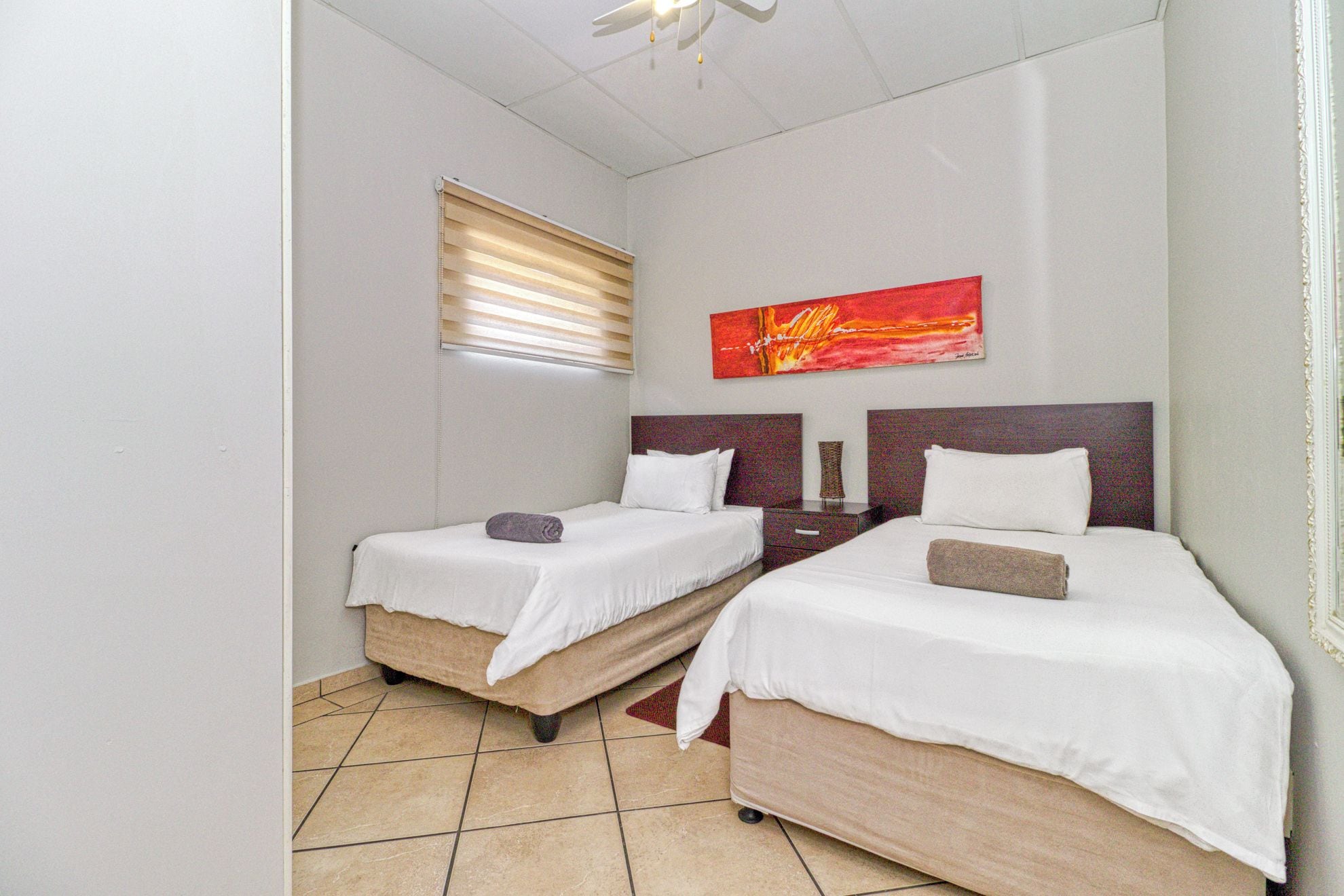 A hotel room with two beds, a window with blinds, a red abstract painting, and a ceiling fan.