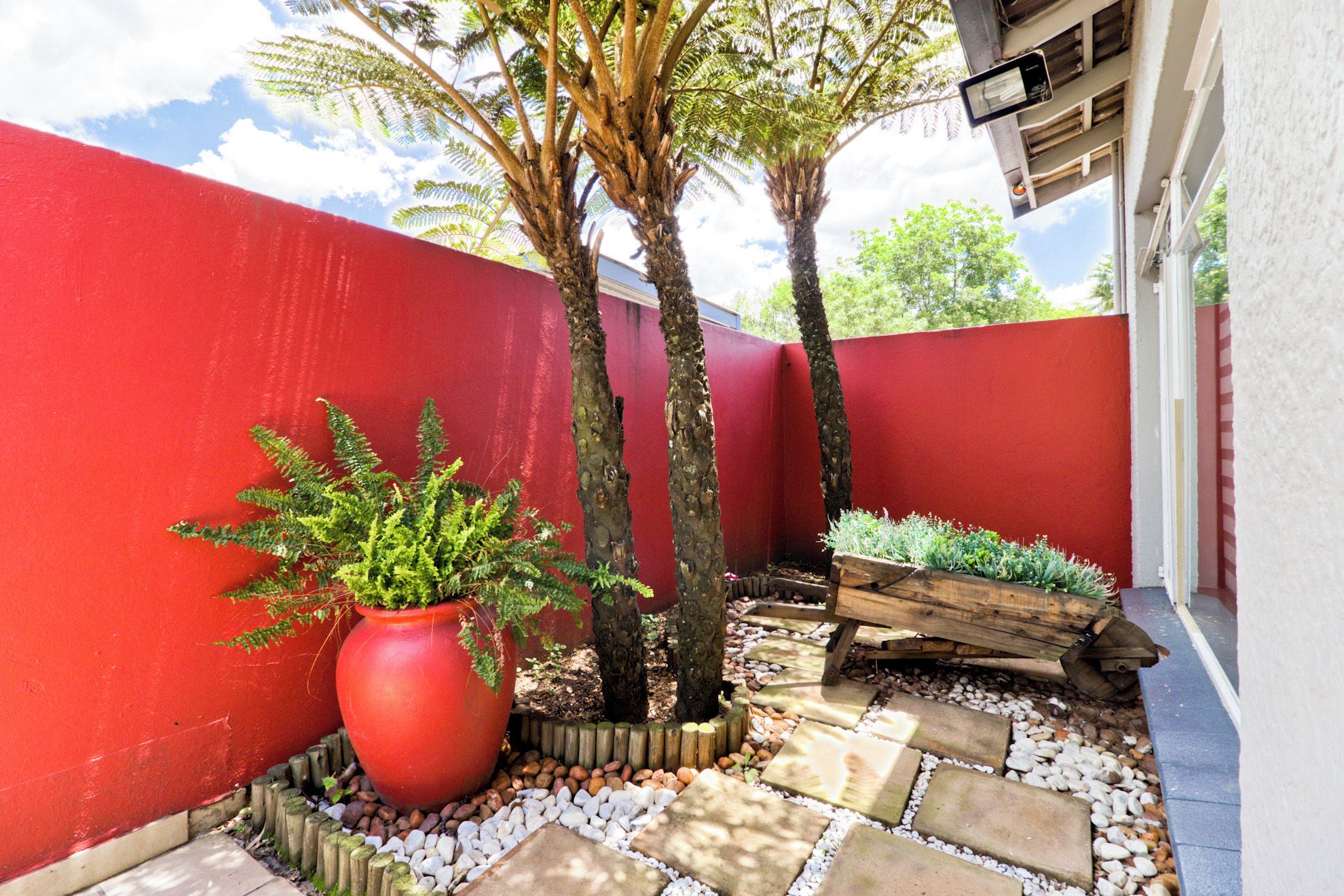 A red-painted exterior wall, a large red planter with greenery, two trees, and a wooden planter bed with plants.