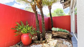 A red-painted exterior wall, a large red planter with greenery, two trees, and a wooden planter bed with plants.