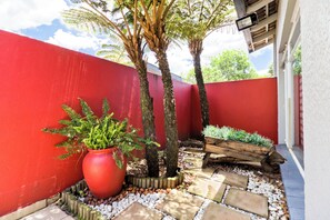 Un mur extérieur peint en rouge, un grand bac à fleurs rouge contenant des plantes vertes, deux arbres et un parterre en bois planté.