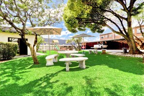 A well-maintained outdoor patio area with stone tables and chairs, surrounded by lush green grass and trees. - Bm Gardens Apartment Hotel (Germiston)