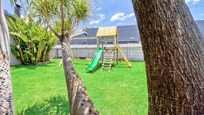 A backyard with a wooden playset, including a slide and climbing area, surrounded by green grass and trees. - Bm Gardens Apartment Hotel (Germiston)