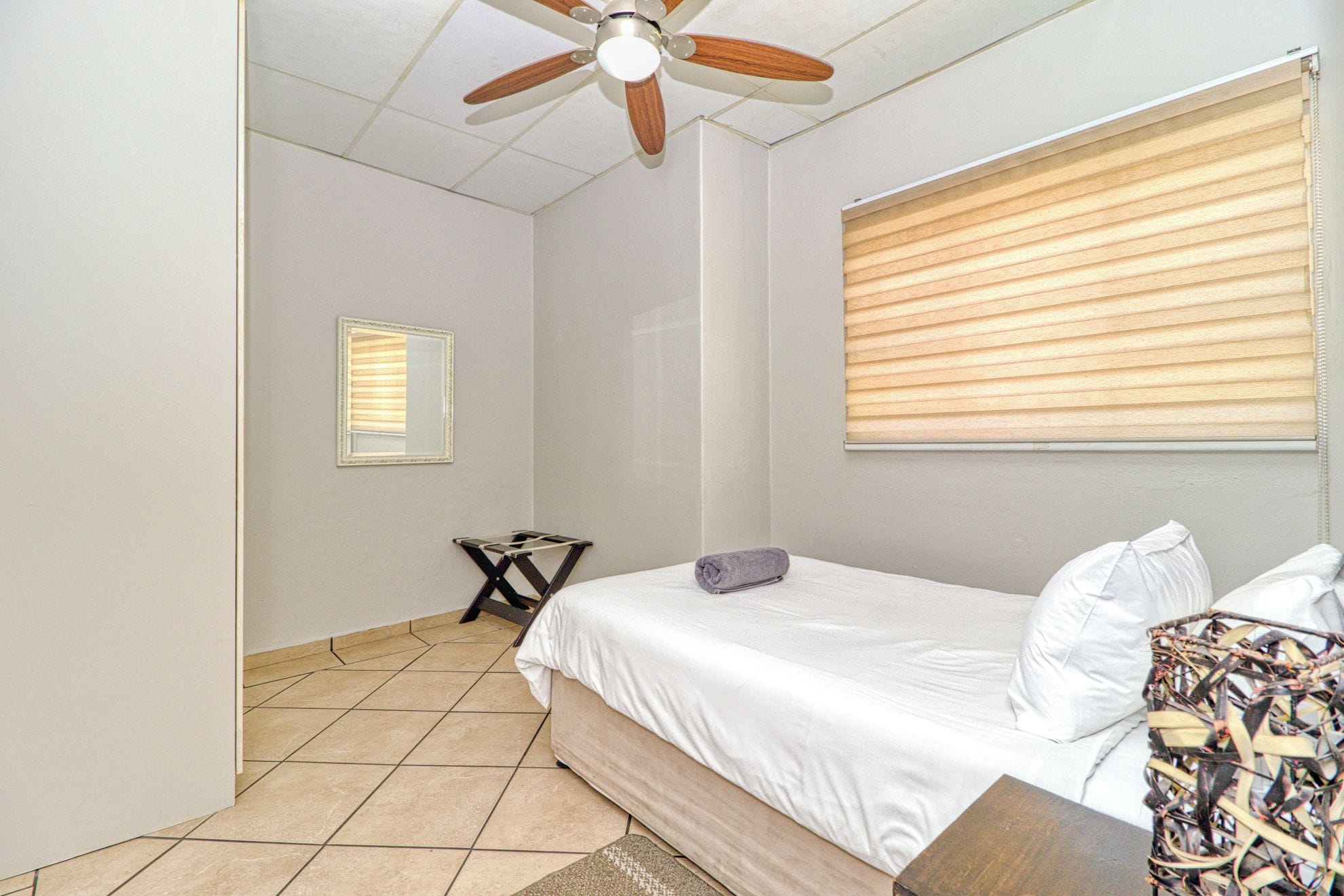 A bedroom with a bed, a ceiling fan, a window with blinds, and a small table with a towel.