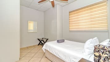 A bedroom with a bed, a ceiling fan, a window with blinds, and a small table with a towel.