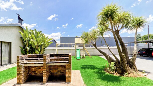 A brick barbecue grill in a grassy yard with a playground and a palm tree. - Bm Gardens Apartment Hotel (Germiston)
