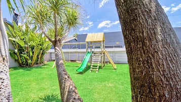 A backyard with a wooden play structure, including a slide and climbing area, surrounded by grass and trees.