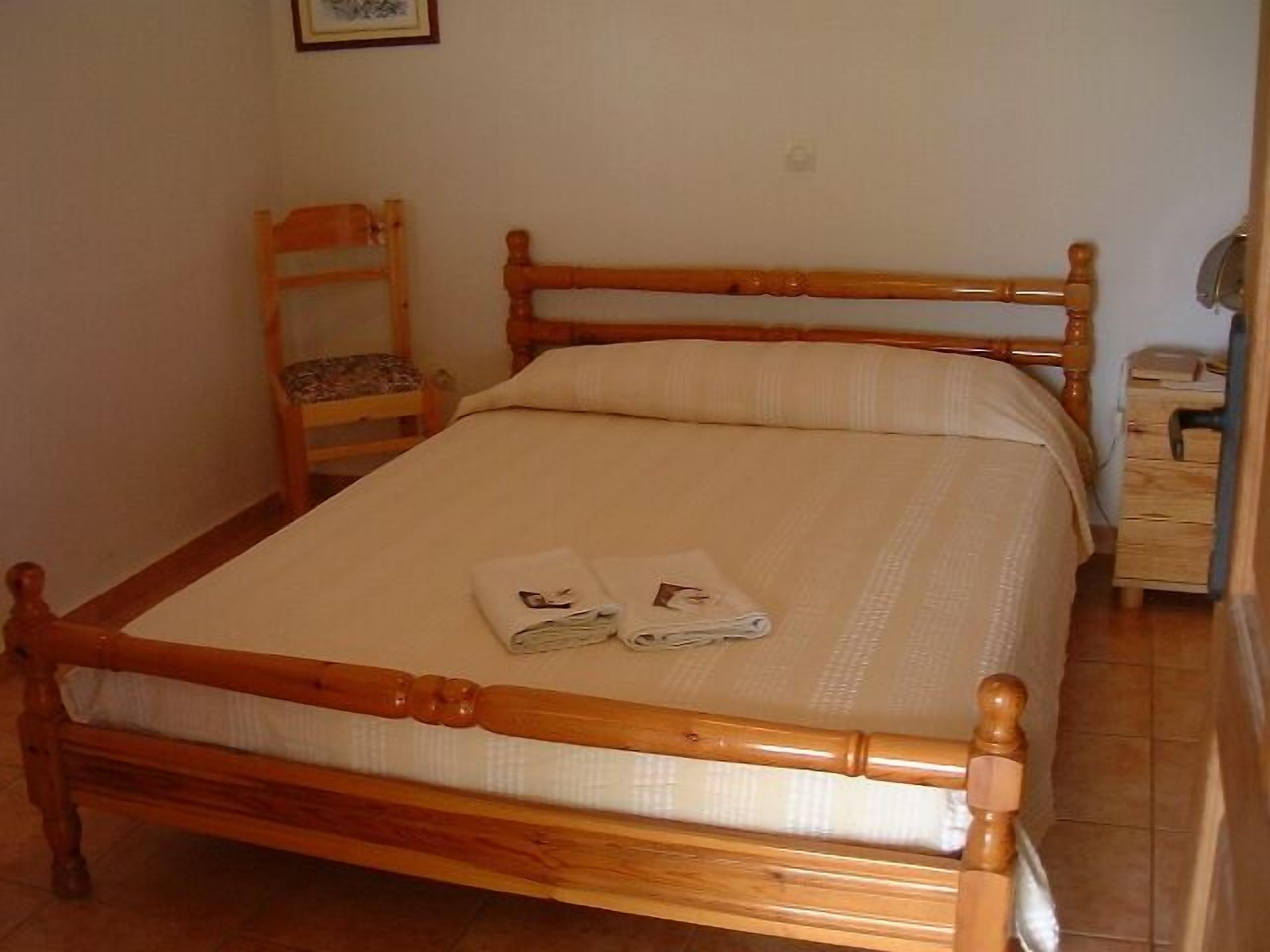 A single bed with a wooden frame and a white mattress, a wooden nightstand with a lamp, and a chair with a cushion.