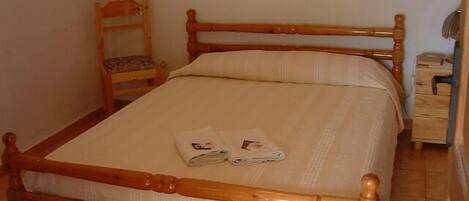 A single bed with a wooden frame and a white mattress, a wooden nightstand with a lamp, and a chair with a cushion.