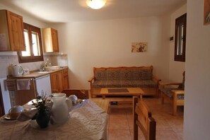 A compact kitchen with wooden cabinets, a dining table with a vase, and a sofa in the living area. - Villa Amalia (Sitia)