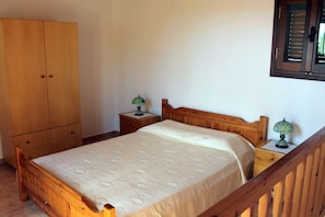 A bedroom with a wooden bed, a wardrobe, two bedside tables with lamps, and a window with shutters. - Villa Amalia (Sitia)