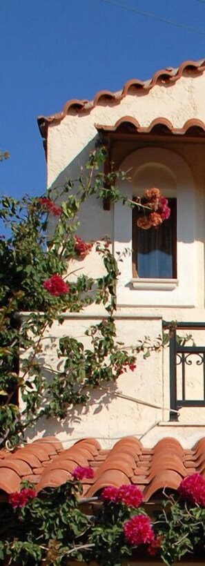 A building with a tiled roof, a window with a curtain, and flowering plants. - Villa Amalia (Sitia)