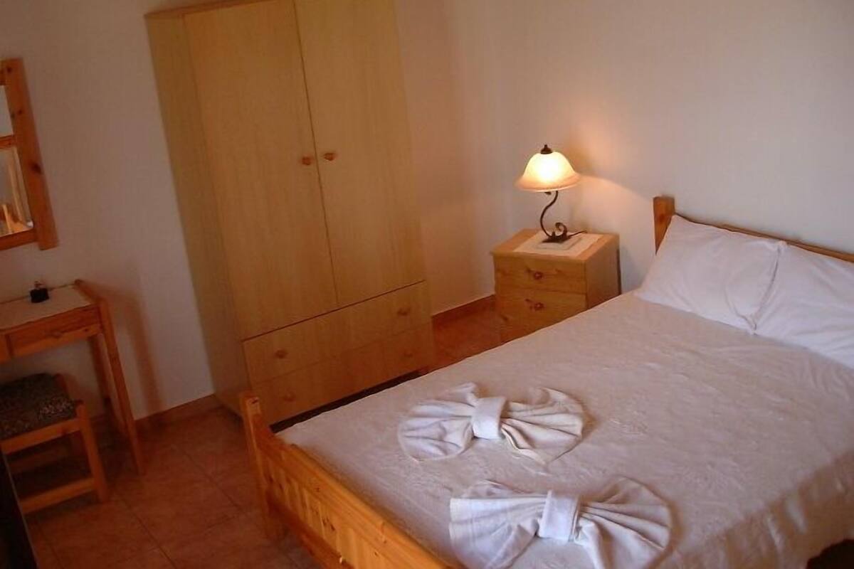 A bedroom with a wooden bed, a nightstand with a lamp, a wooden wardrobe, and a small wooden desk with a chair.