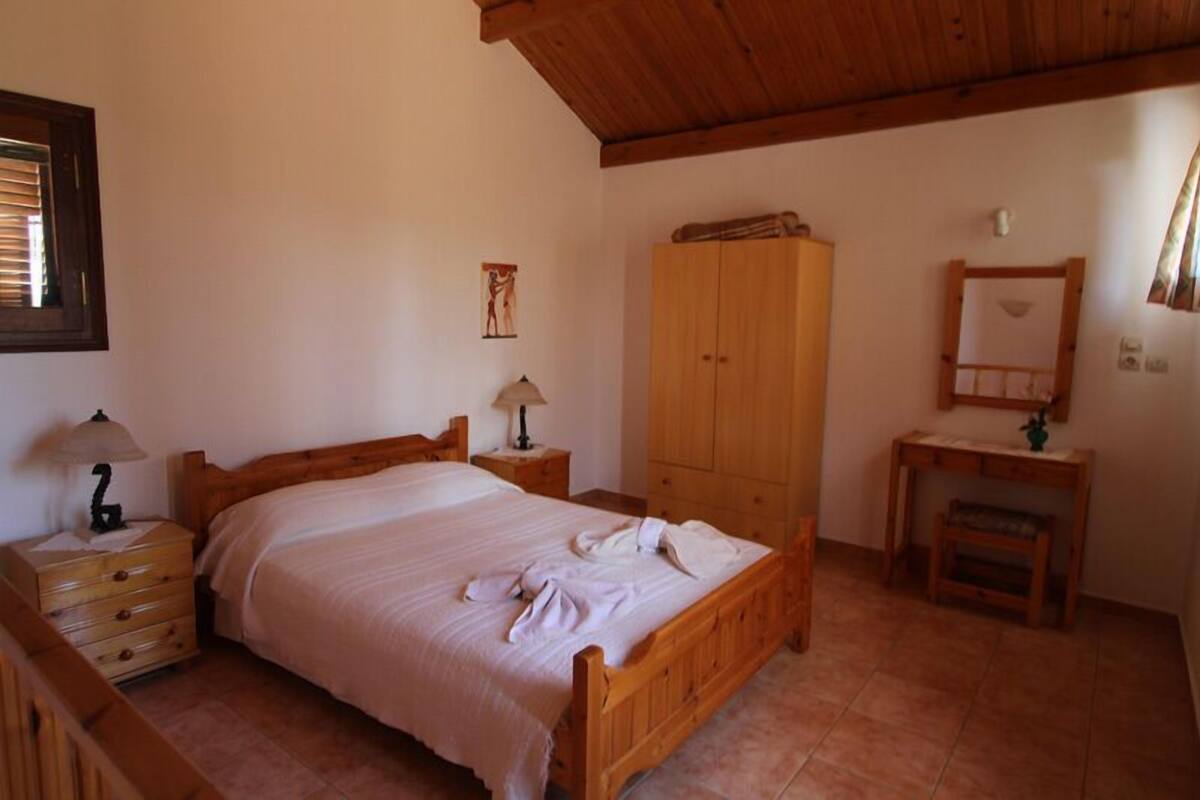 A bedroom with a wooden bed, a nightstand with a lamp, a wooden wardrobe, and a small table with a mirror.