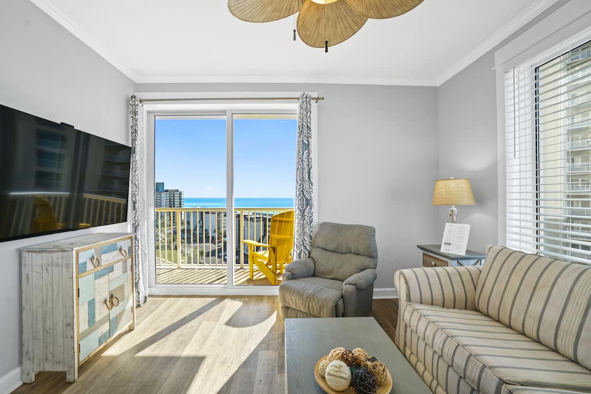 A living room with a large TV, a sofa, a chair, a coffee table, and a balcony with a view of the ocean.