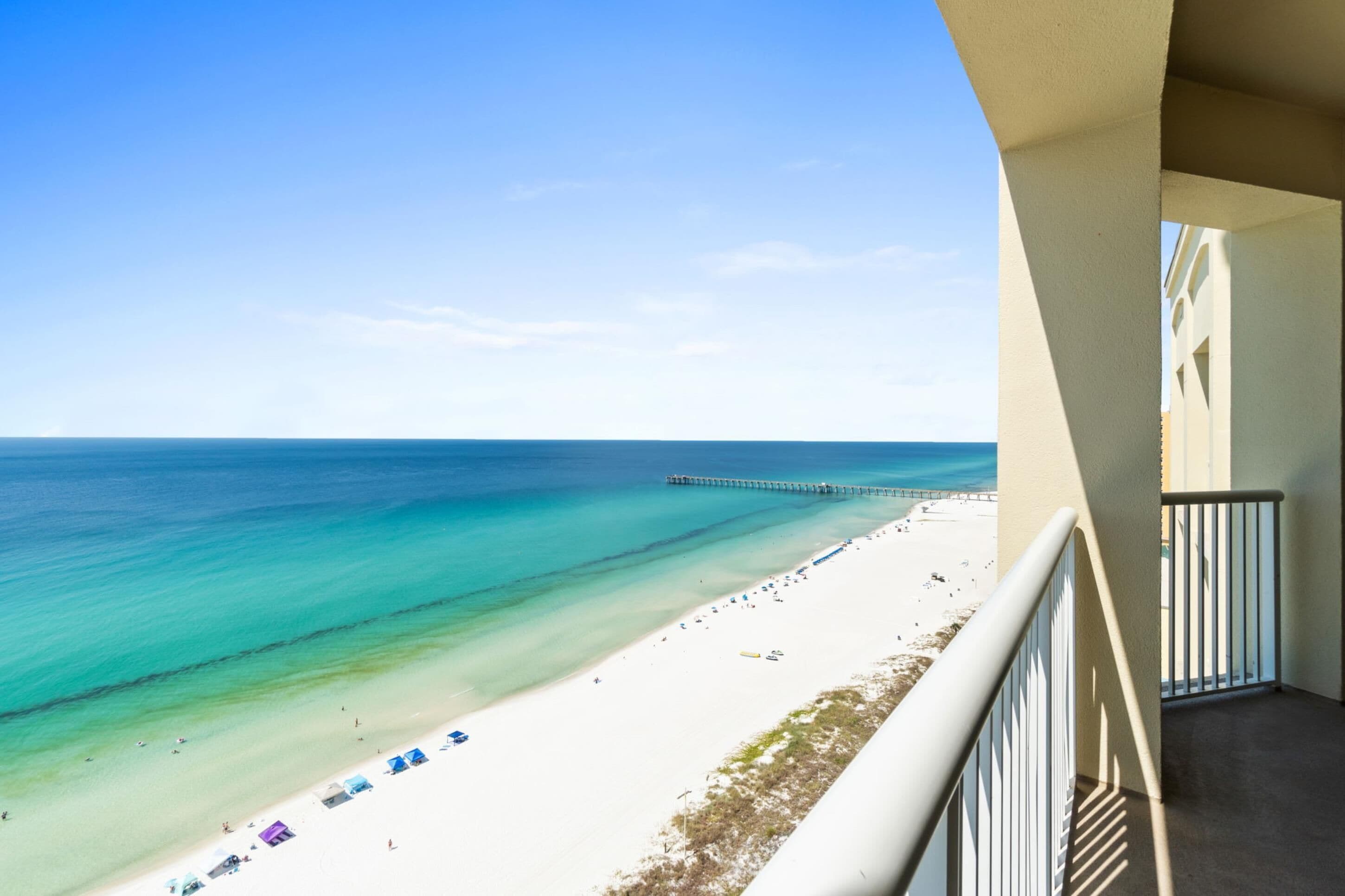 Blick auf den Strand vom Balkon, mit blauem Himmel, einem Steg in der Ferne und einem sandigen Ufer mit Strandsonnenschirmen.