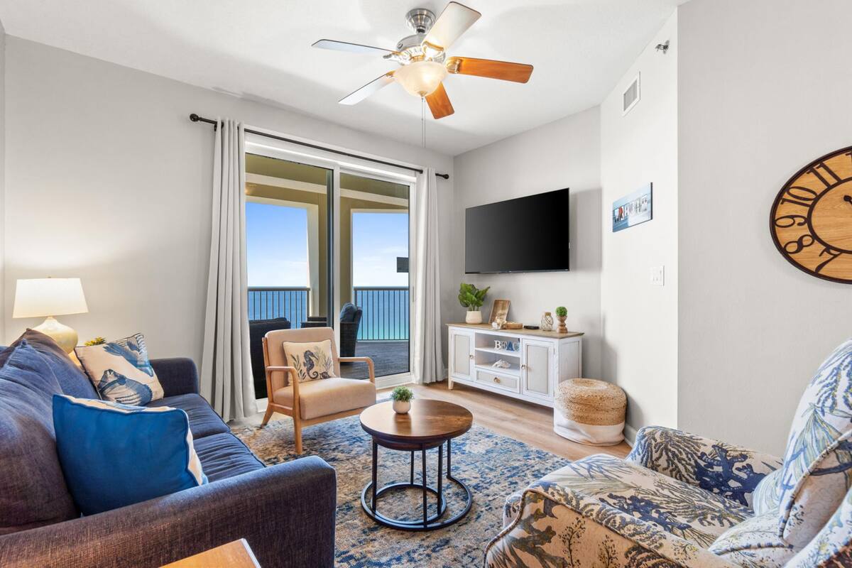A living room with a sofa, armchair, and a coffee table. There is a television mounted on the wall and a ceiling fan.