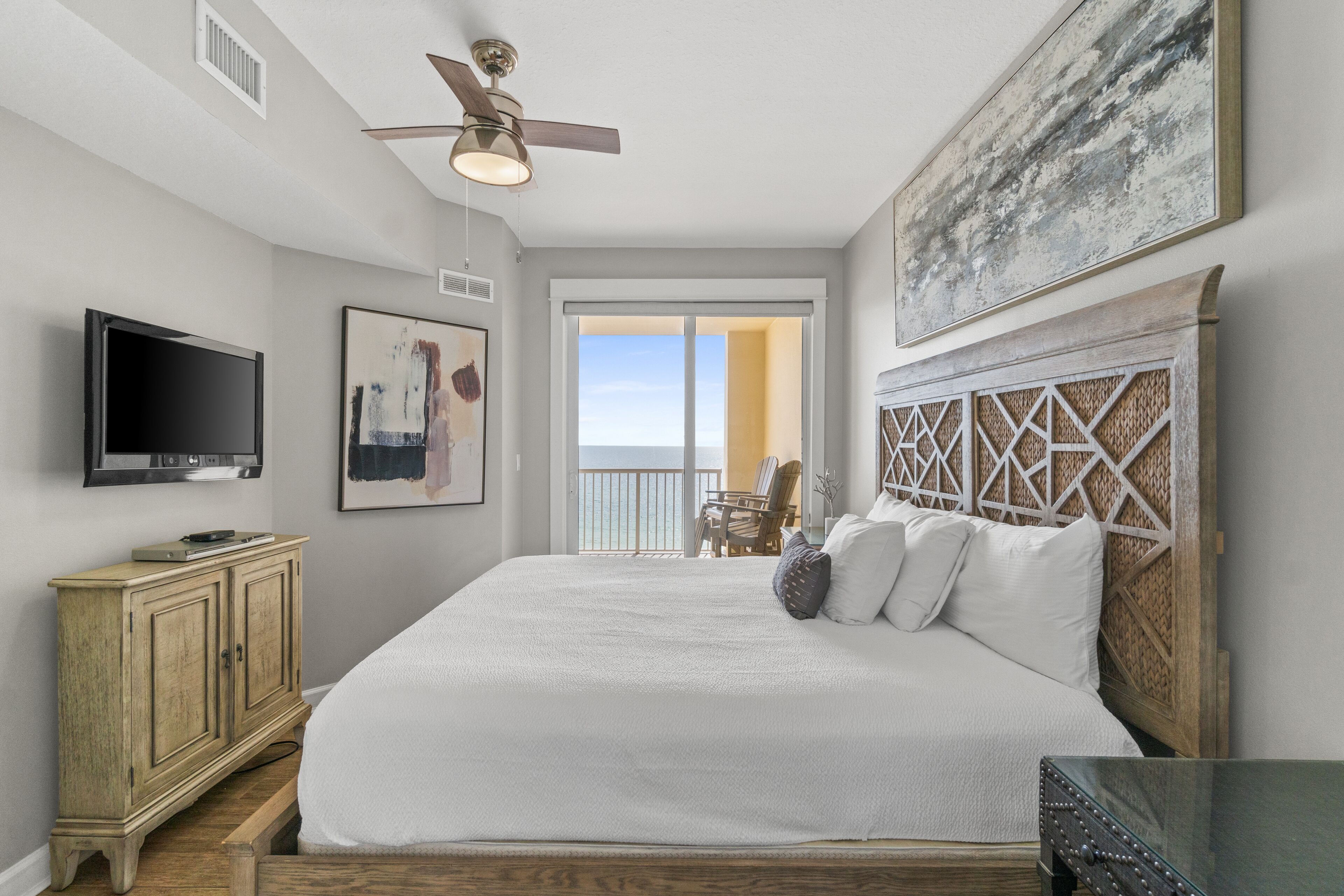 A bedroom with a large bed, a ceiling fan, a TV, and a view of the outdoors.