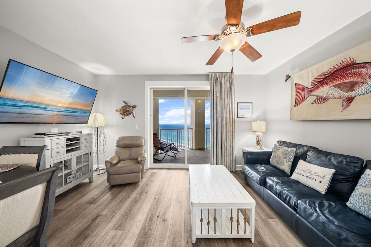 A living room with a ceiling fan, a TV, a couch, a coffee table, and a view of the ocean through a sliding door.