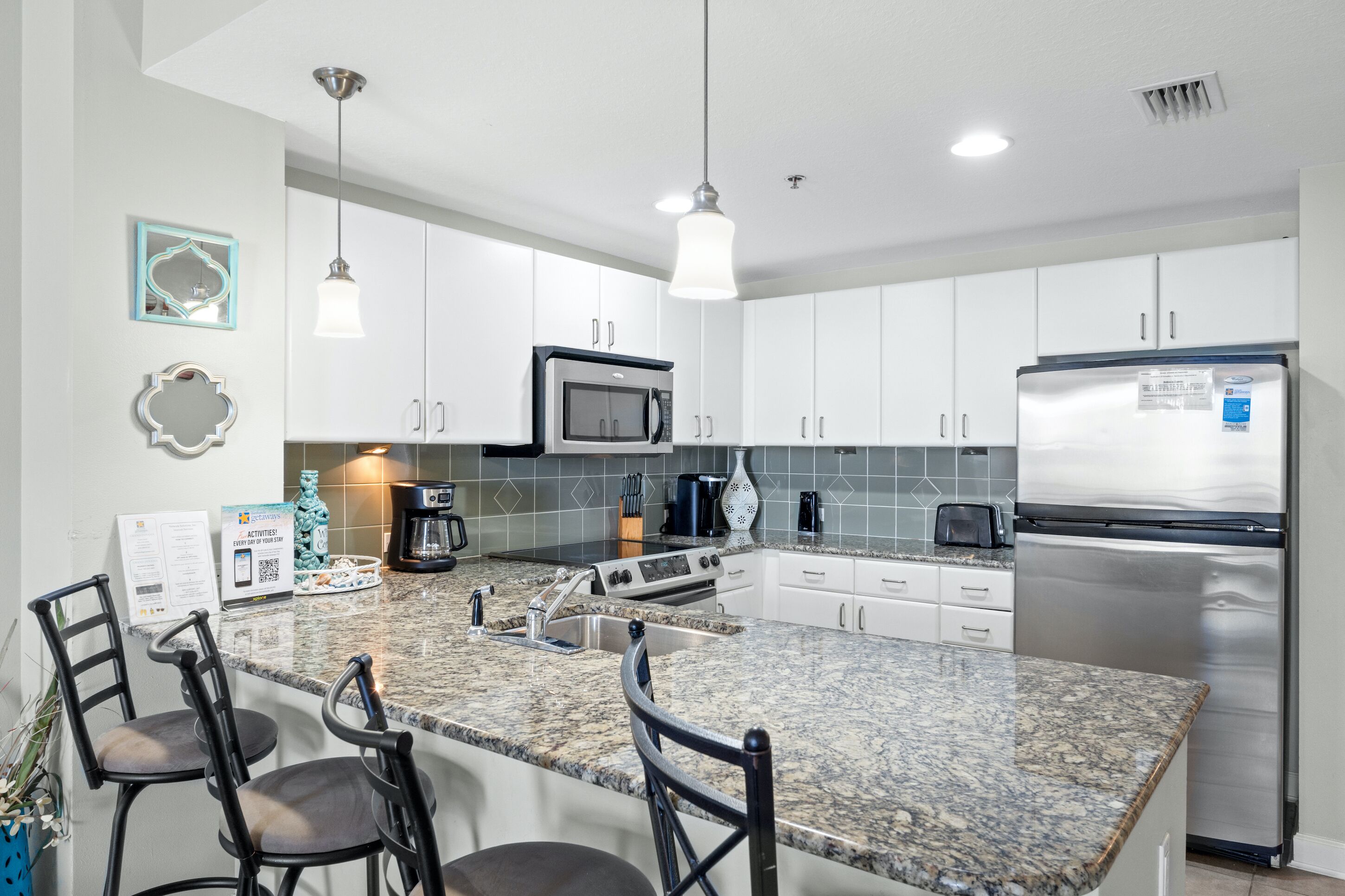 A modern kitchen with white cabinets, a stainless steel refrigerator, a microwave, and a granite countertop. There are four bar stools and a coffee maker on the counter.