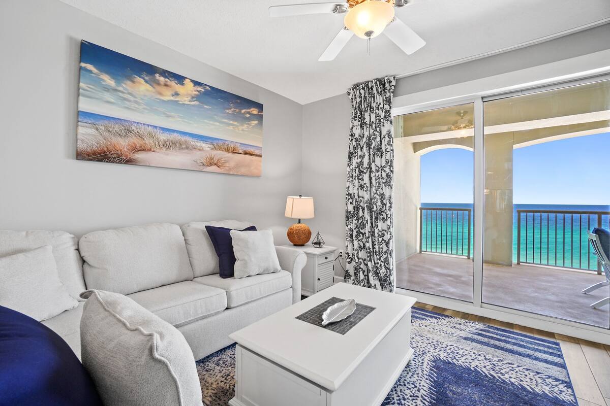 A modern living room with a large window, a ceiling fan, a sofa, a coffee table, and a painting of a beach scene.