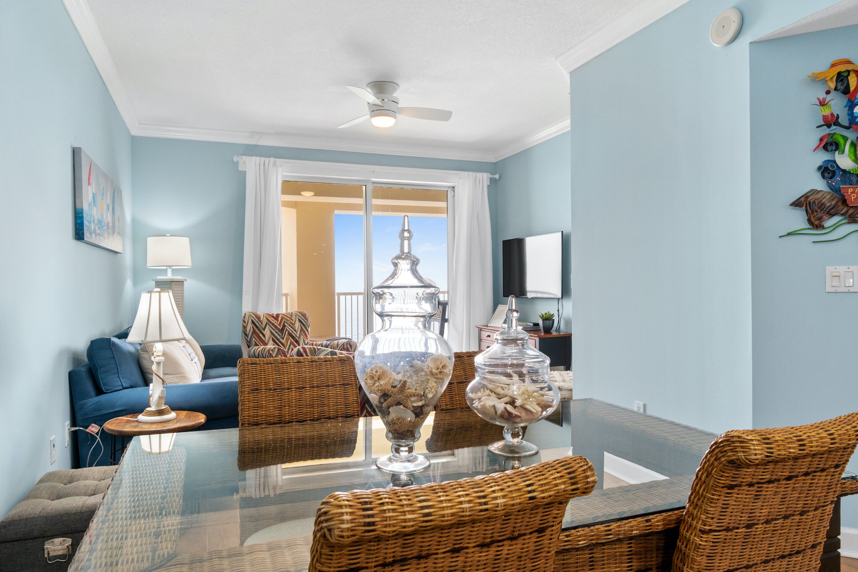 Una sala de estar con una mesa de comedor de cristal, sillas de mimbre, un sofá azul, una lámpara blanca y una decoración de pájaros en la pared.