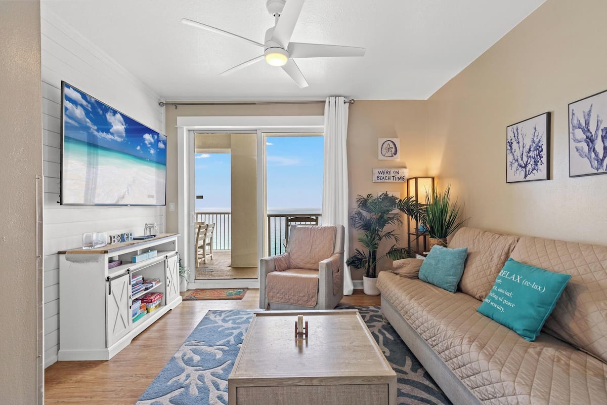 A living room with a ceiling fan, a sofa with teal pillows, a coffee table, a bookshelf, and a view to the outside.