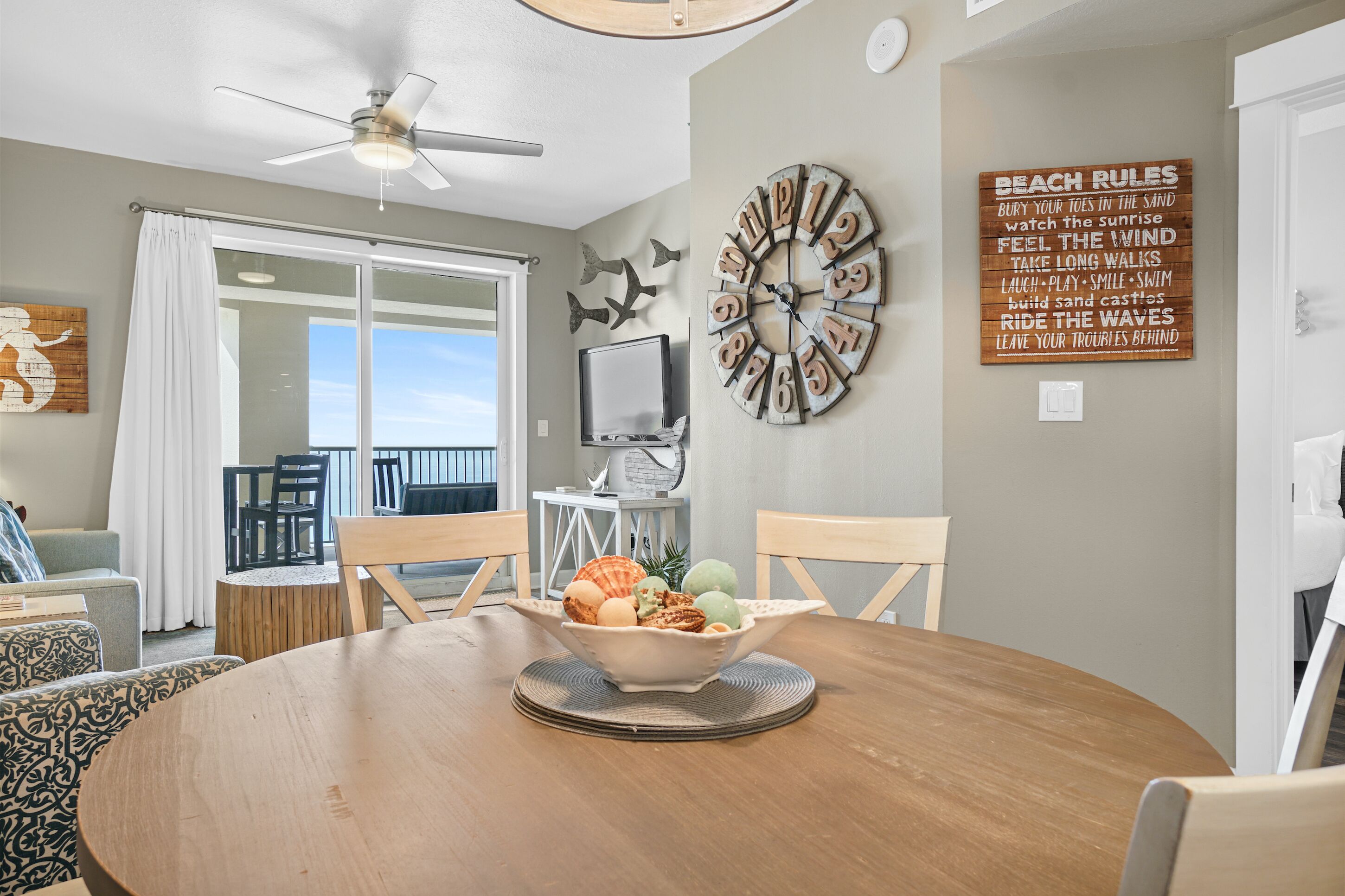 A dining area with a round table, chairs, a bowl of fruit, a clock, and a beach-themed wall decoration.