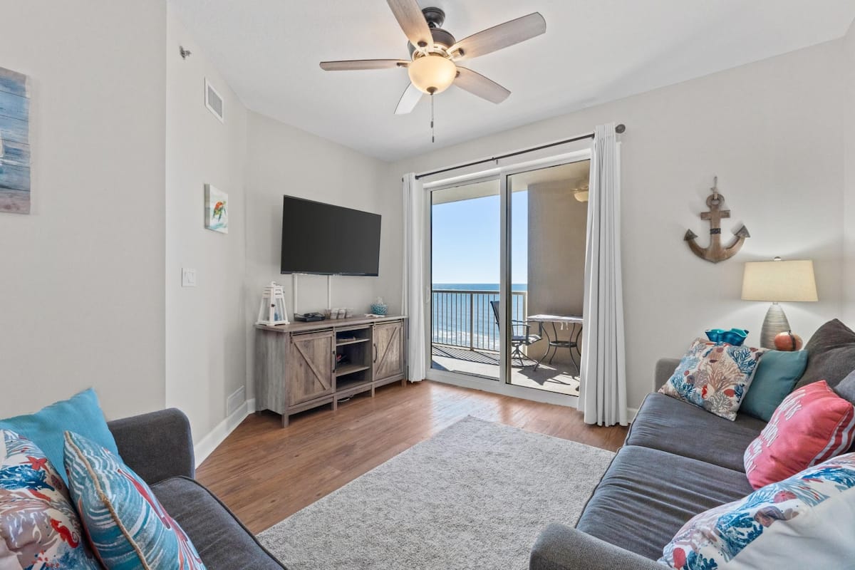 A living room with a ceiling fan, a flat-screen TV, a sliding glass door to a balcony, and a sofa with decorative pillows.