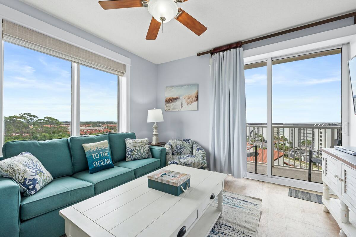 A modern living room with a teal sofa, a white coffee table, and a view of the city through large windows.