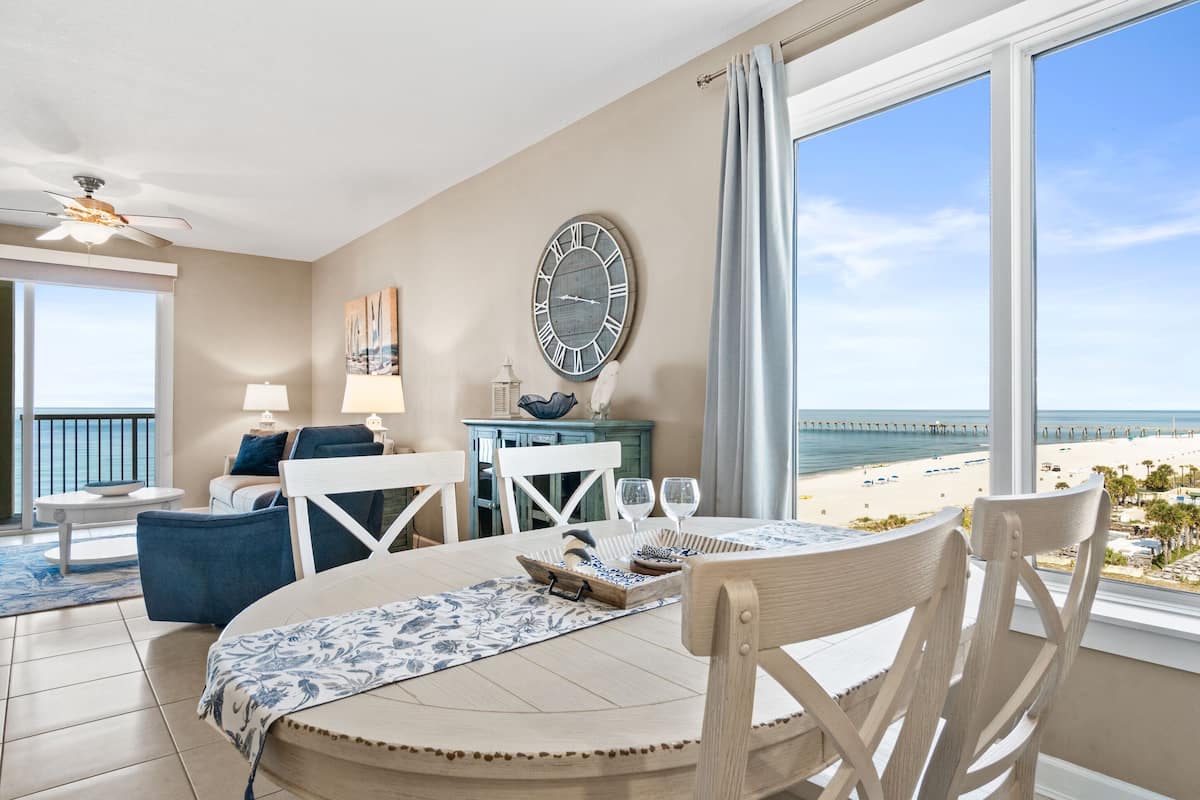 A dining area with a view of the beach, a clock on the wall, and a ceiling fan.