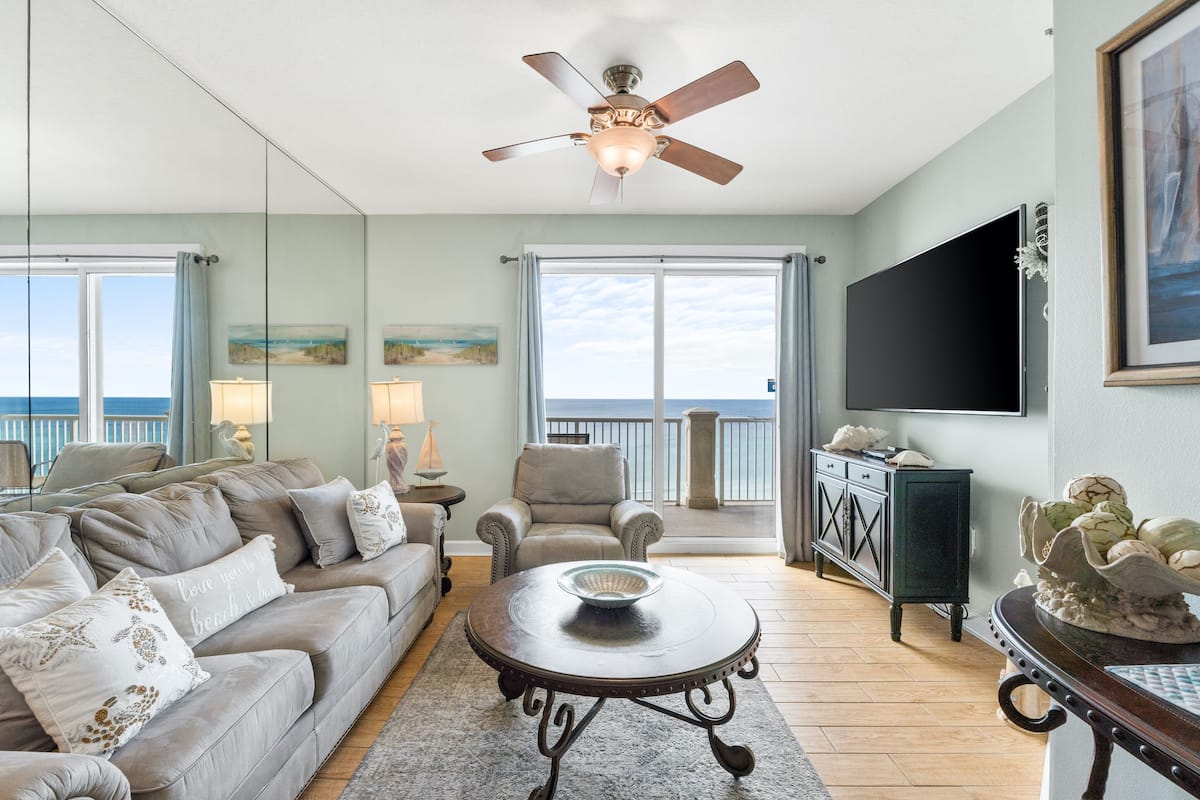 A living room with a sofa, armchairs, a coffee table, a ceiling fan, a TV, and a view of the ocean through large windows.