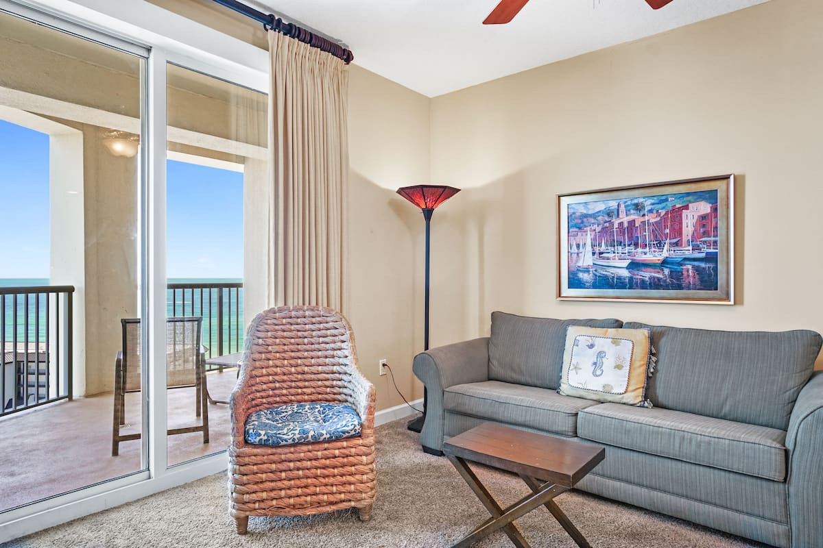 A living room with a wicker chair, a gray sofa, a painting of a harbor, and a view of the ocean through a sliding glass door.