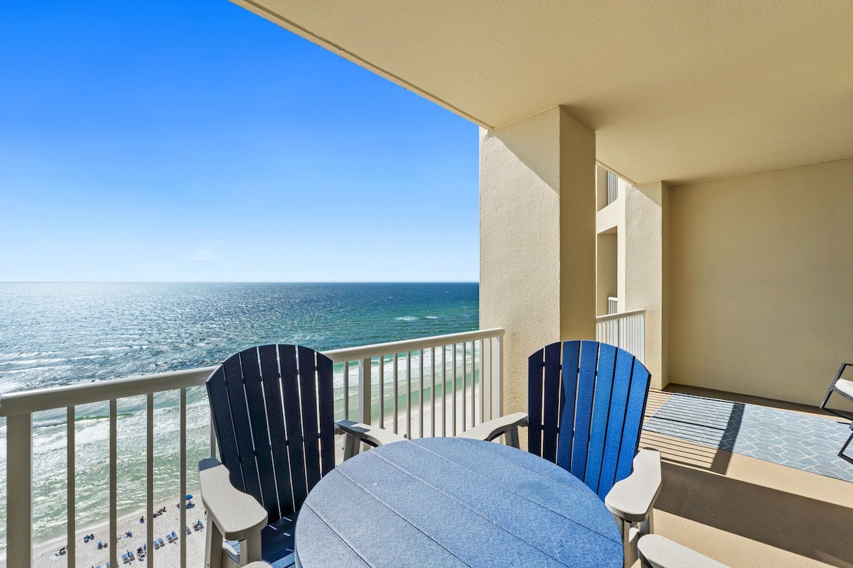 A balcony with two Adirondack chairs and a round table, overlooking a beach and the ocean.