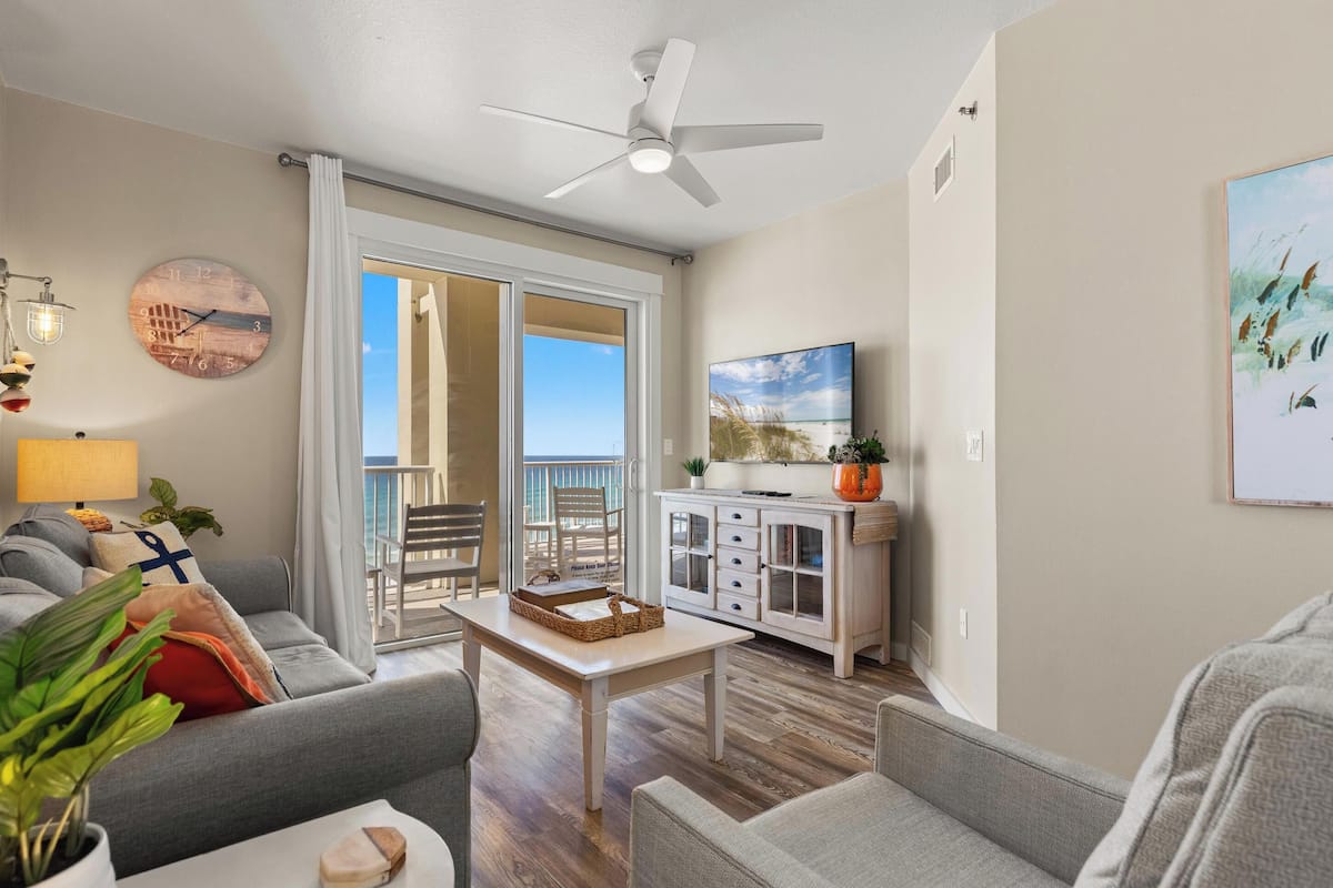 A living room with a sofa, a coffee table, a TV, and a balcony with a view.