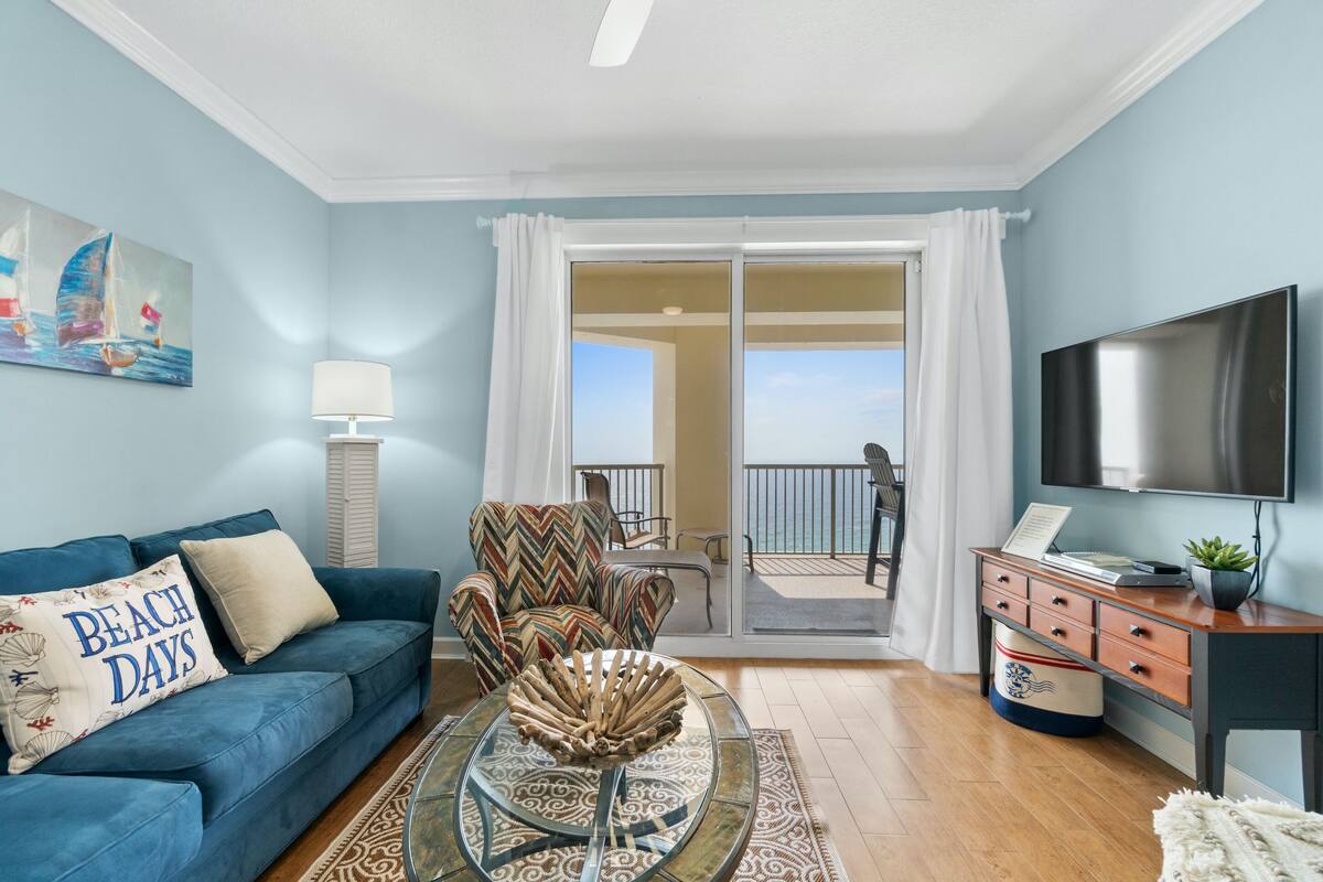 A living room with a blue sofa, a patterned armchair, a glass coffee table, a flat-screen TV, and a view of the balcony.