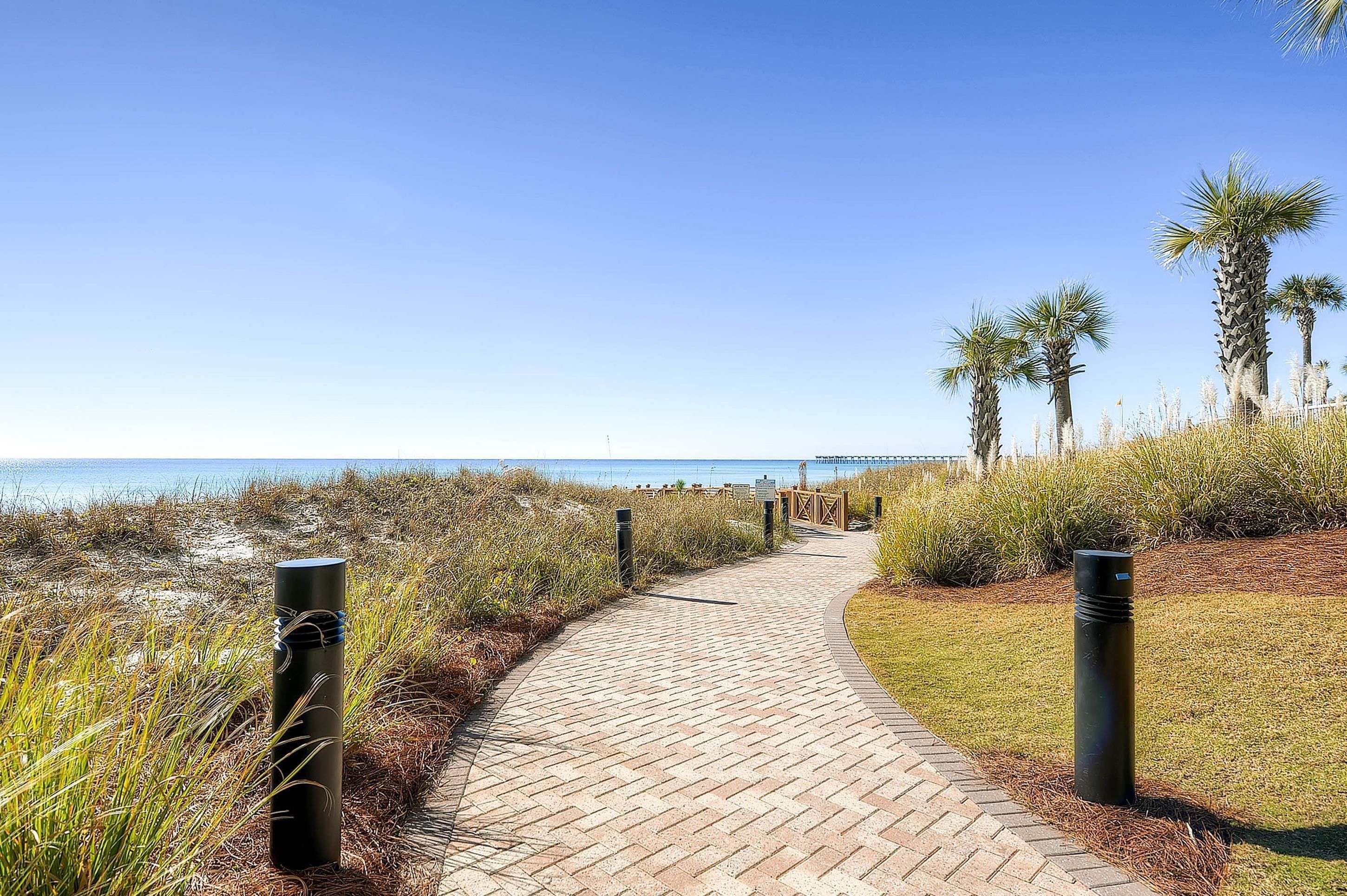 Un sendero pavimentado que conduce a una playa, bordeado por césped y palmeras.