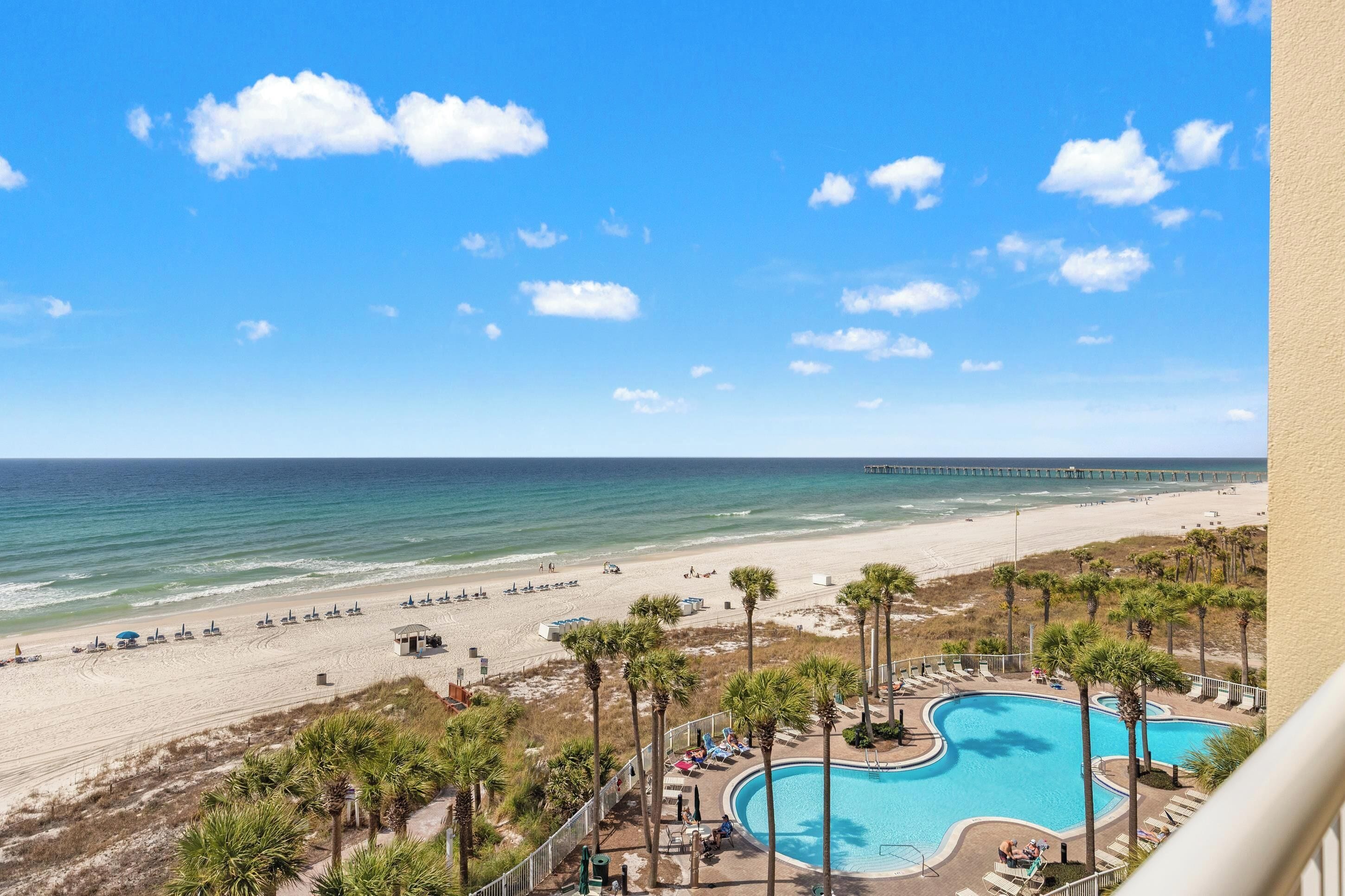 A beach view with a swimming pool, palm trees, and a pier in the distance.
